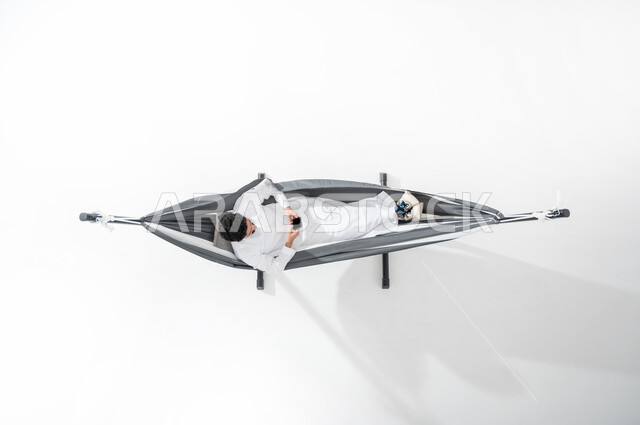 استخدام الأجهزة الالكترونية الحديثة, بورتريه علوي عامودي لشاب عربي خليجي سعودي مبتسم يرتدي الثوب التقليدي يجلس على أرجوحة قماشية معلقة ويمسك بيده الهاتف المحمول, عرض شاشة الجوال, الترويح عن النفس, خلفية بيضاء