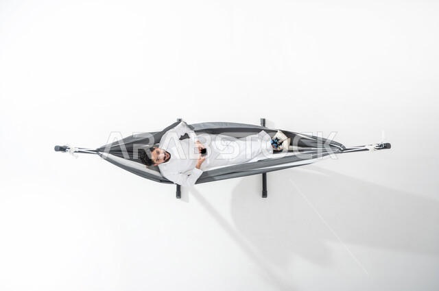 الأجهزة الالكترونية الحديثة, بورتريه علوي عامودي لشاب عربي خليجي سعودي مبتسم يرتدي الثوب التقليدي يجلس على أرجوحة قماشية معلقة ويمسك بيده الهاتف المحمول, عرض شاشة الجوال سوداء فارغة, النظر إلى الأعلى, الترويح عن النفس, خلفية بيضاء