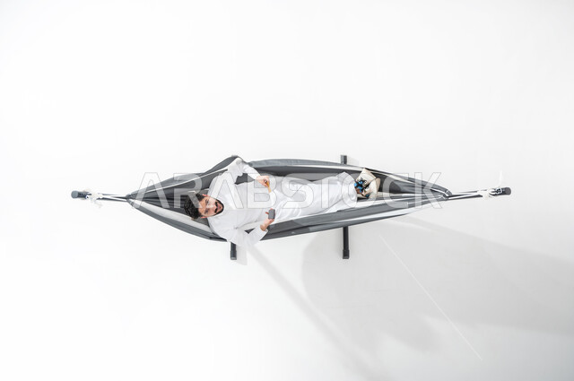 الاستفادة من التكنولوجيا المتطورة والتقنيات الحديثة, بورتريه علوي عامودي لشاب عربي خليجي سعودي مبتسم يرتدي الثوب التقليدي يجلس على أرجوحة قماشية معلقة ويقوم بالدفع الالكتروني بواسطة الهاتف المحمول, استخدام بطاقة الصراف, سهولة الدفع عبر الحساب البنكي, خلفية بيضاء