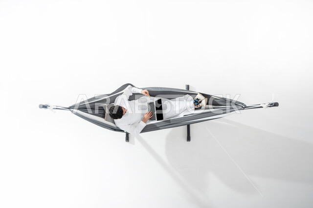 Using advanced technology and modern techniques, a vertical overhead portrait of a smiling Saudi Gulf Arab young man wearing a traditional dress, sitting on a hanging cloth hammock and making electronic payments using a laptop, using an ATM card, ease of paying via a bank account, white background