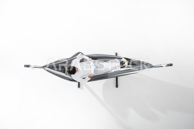 ممارسة هواية قراءة المجلات، استغلال وقت الاستراحة بالمطالعة, بورتريه علوي عامودي لشاب مبتسم عربي خليجي سعودي يرتدي الثوب التقليدي يجلس على أرجوحة قماشية معلقة ويمسك بيده مجلة ثقافية, النظر إلى الأعلى، قضاء الوقت بعيداً عن التكنولوجيا