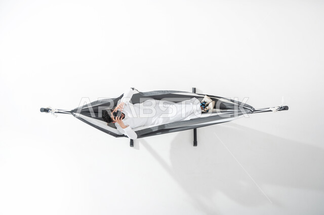 ممارسة الهوايات في أوقات الاستراحة, بورتريه عامودي علوي لشاب عربي خليجي سعودي يرتدي الثوب التقليدي ويستلقي على أرجوحة قماشية معلقة يمسك بيده آلة التصوير الرقمية, السعودية, التقاط الصور الفوتوغرفية من خلال الكاميرا, تقريب الأجسام البعيدة, قضاء الوقت مع الذات