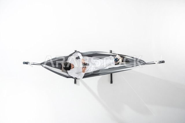 ممارسة الهوايات في أوقات الاستراحة, بورتريه عامودي علوي لشاب عربي خليجي سعودي يرتدي الثوب التقليدي ويستلقي على أرجوحة قماشية معلقة يمسك بيده آلة التصوير الرقمية, السعودية, التقاط الصور الفوتوغرفية من خلال الكاميرا, تقريب الأجسام البعيدة, قضاء الوقت مع الذات