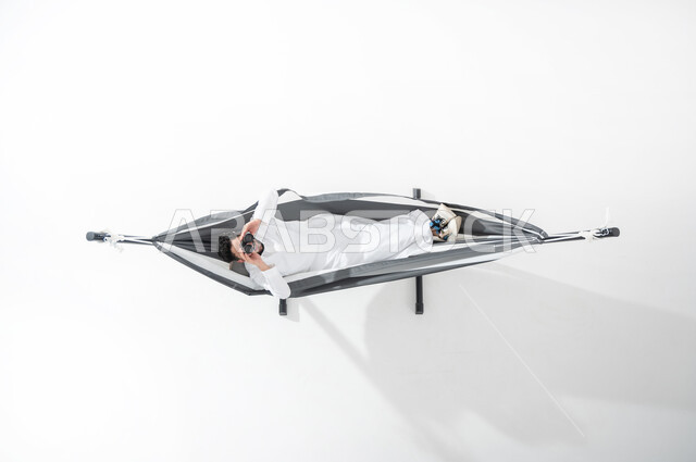 الاسترخاء والهدوء خلال الاستراحة, بورتريه عامودي علوي لشاب عربي خليجي سعودي يرتدي الثوب التقليدي ويستلقي على أرجوحة قماشية معلقة، النظر من خلال المنظار المكبر بعدستين, تقريب الأشياء البعيدة, الإشارة إلى رؤية جسم بعيد, رؤية مستقبلية سعودية, متعة قضاء الوقت مع الذات