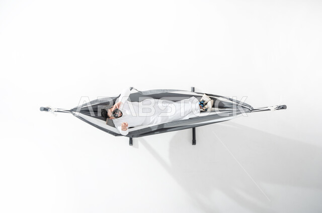 الاسترخاء والاستمتاع بأوقات الاستراحة, بورتريه عامودي علوي لشاب عربي خليجي سعودي يرتدي الثوب التقليدي ويستلقي على أرجوحة قماشية معلقة، النظر من خلال المنظار المكبر, تقريب الأشياء البعيدة, الإشارة إلى رؤية جسم بعيد, رؤية مستقبلية سعودية, متعة قضاء الوقت مع الذات