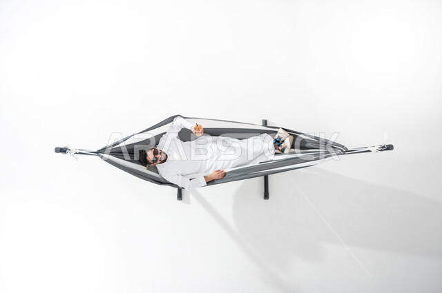 قضاء أوقات ممتعة في الاستراحة , بورتريه عامودي علوي لشاب عربي خليجي سعودي يرتدي الثوب التقليدي مع نظارات شمسية يقوم بالاستلقاء على ارجوحة قماشية معلقة أثناء شربه للعصير الطازج , الاسترخاء و الراحة والسكون , الاستمتاع بالمشروبات الصيفية الباردة ,خلفية بيضاء