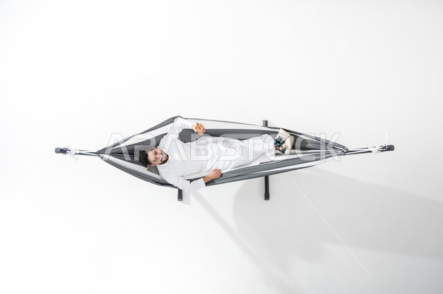 قضاء الوقت بالاسترخاء و السكون , بورتريه عامودي علوي لشاب عربي خليجي سعودي يرتدي الثوب التقليدي يقوم بالاستلقاء على أرجوحة قماشية معلقة  يشرب عصير منعش , النظر إلى الكاميرا بإيماءات السعادة و السرور , الاستمتاع بالمشروبات الصيفية الباردة, خلفية بيضاء