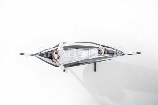 Wearing 3D virtual reality glasses, a vertical overhead portrait of a Saudi Gulf Arab young man wearing a traditional dress, lying on a hanging cloth hammock and trying to focus and integrate, gestures indicating interaction, pleasure and amusement, white background