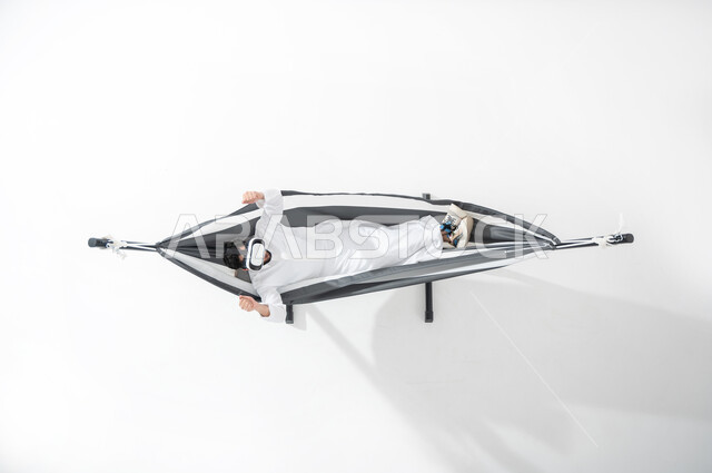 المتعة و التسلية في العالم الافتراضي , بورتريه علوي عامودي لشاب عربي خليجي سعودي يرتدي الثوب التقليدي يستلقي على ارجوحة قماشية معلقة يقوم بوضع نظارات الواقع الافتراضي الفي ار بإيماءات تدل على الاندماج و التشجيع  , خلفية بيضاء