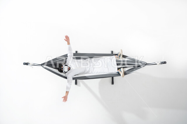 تجربة نظارات الواقع الافتراضي , بورتريه علوي عامودي لشاب عربي خليجي سعودي يرتدي الثوب التقليدي يقوم بالاستلقاء على أرجوحة قماشية معلقة و ارتداء نظارة الفي ار , إيماءات تدل على التفاعل و المتعة و التسلية , خلفية بيضاء