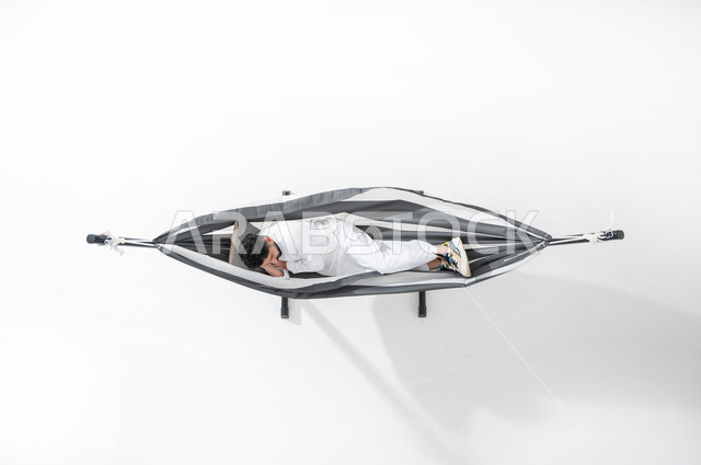 وقت الاستراحة و الاسترخاء و السكون , بورتريه علوي عامودي لشاب عربي خليجي سعودي يرتدي الثوب التقليدي يقوم بالاستلقاء على أرجوحة قماشية معلقة ليأخذ قسط من النوم و الراحة بعد العمل , خلفية بيضاء