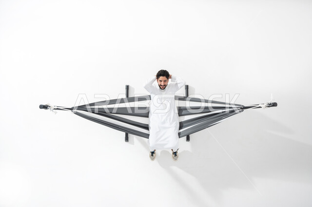 الحاجة إلى الراحة و الهدوء , بورتريه علوي عامودي لشاب عربي خليجي سعودي يرتدي الثوب التقليدي  يجلس على أرجوحة قماشية معلقة يضع يديه خلف رأسه , النظر إلى الكاميرا بإيماءات الاسترخاء و السرور ,  قضاء الأوقات الممتعة بالعطلة , خلفية بيضاء