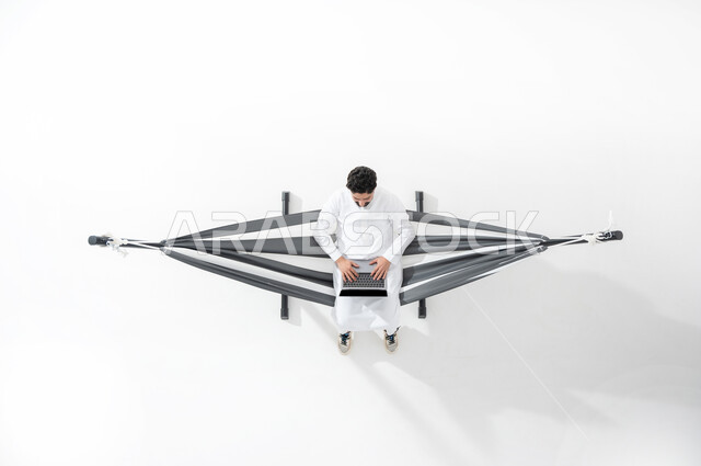 استخدام أجهزة الحواسيب الحديثة و المتطورة في متابعة الأعمال و المشاريع , بورتريه علوي عامودي لشاب عربي خليجي سعودي يرتدي الثوب التقليدي يجلس على أرجوحة قماشية معلقة يقوم بالكتابة على لوحة المفاتيح لإنجاز المهام عن بعد , خلفية بيضاء