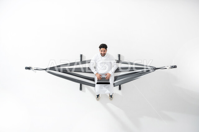 Using advanced technologies, a vertical overhead portrait of a Saudi Gulf Arab young man wearing a traditional dress, sitting on a hanging cloth swing, doing his work on the computer, looking up with gestures of happiness and pleasure, white background..