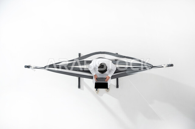 تصفح الانترنت و وسائل التواصل الاجتماعي باستخدام التقنيات الحديثة و المتطورة , انجاز المهام عن بعد ,بورتريه علوي عامودي لشاب عربي خليجي سعودي يرتدي الثوب التقليدي يجلس على أرجوحة قماشية معلقة يقوم بالعمل على جهاز الحاسوب المحمول , خلفية بيضاء