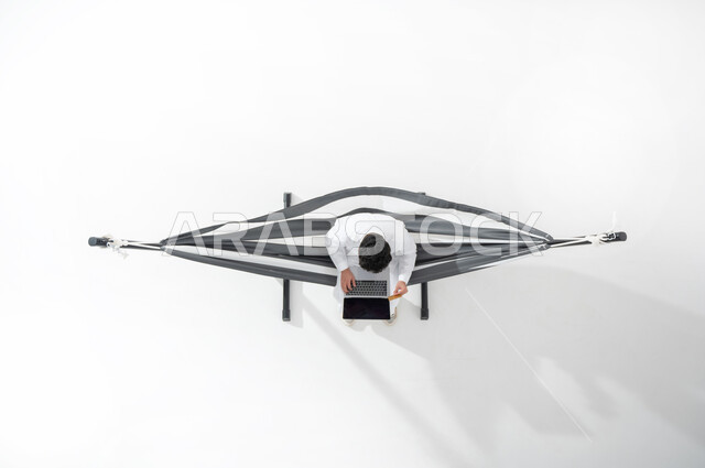 استخدام التقنية و التكنولوجيا الحديثة , انجاز مهام العمل عن بعد,  بورتريه علوي عامودي لشاب عربي خليجي سعودي يرتدي الثوب التقليدي يجلس على أرجوحة قماشية معلقة يقوم بعمله على جهاز الحاسوب , عرض شاشة سوداء  , خلفية بيضاء