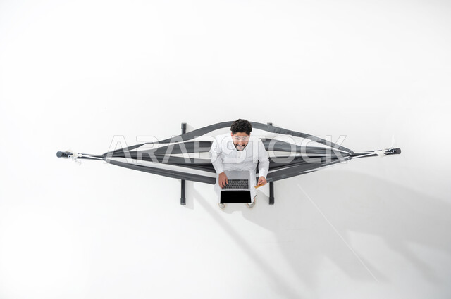 انجاز مهام العمل عن بعد , استخدام التقنية و التكنولوجيا الحديثة , بورتريه علوي عامودي لشاب عربي خليجي سعودي يرتدي الثوب التقليدي يجلس على أرجوحة قماشية معلقة , عرض شاشة سوداء فارغة عبر جهاز الحاسوب , خلفية بيضاء