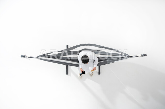 استخدام الأجهزة الالكترونية الحديثة , بورتريه علوي عامودي لشاب عربي خليجي سعودي يرتدي الثوب التقليدي يجلس على أرجوحة قماشية معلقة  يمسك هاتفه المحمول بيده  , عرض شاشة سوداء فارغة عبر الجوال , خلفية بيضاء