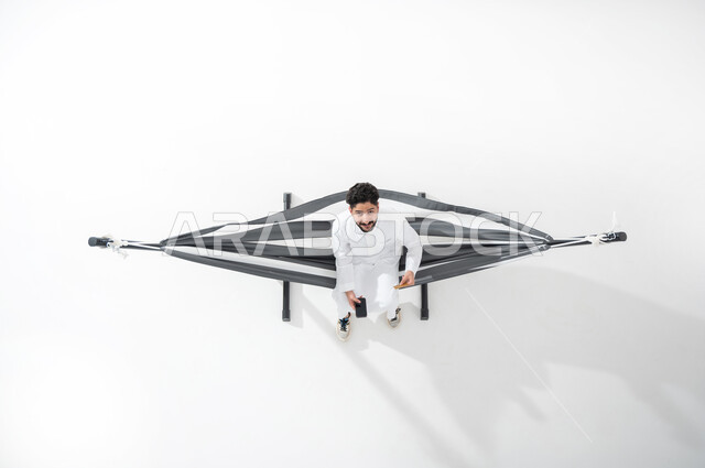استخدام التقنيات الحديثة و المعاصرة للتواصل , بورتريه علوي عامودي لشاب عربي خليجي سعودي يرتدي الثوب التقليدي يجلس على أرجوحة قماشية معلقة  يمسك هاتفه بيده  ينظر إلى الأعلى بإيماءات السعادة و السرور , عرض شاشة سوداء فارغة عبر الجوال ,خلفية بيضاء