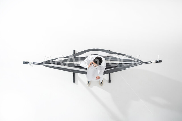 Communicating with family and friends, using advanced electronic devices, vertical overhead portrait of a Saudi Gulf Arab young man wearing a traditional dress, sitting on a hanging cloth hammock and holding a mobile phone in his hand, making a call via mobile phone, white background