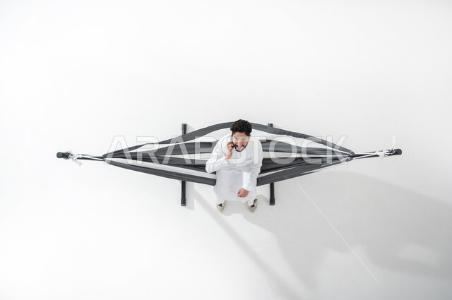 Mobile phone call, vertical portrait of a Saudi Gulf Arab young man wearing a traditional dress sitting on a hanging cloth hammock, holding the mobile phone in his hand, a man making a phone call, white background