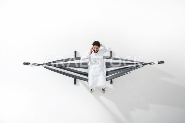 إجراء مكالمة هاتفية , بورتريه علوي عامودي لشاب عربي خليجي سعودي يرتدي الثوب التقليدي يجلس على أرجوحة قماشية معلقة يقوم بإجراء مكالمة هاتفية عبر الجوال , التواصل مع الأهل و الأصدقاء , خلفية بيضاء