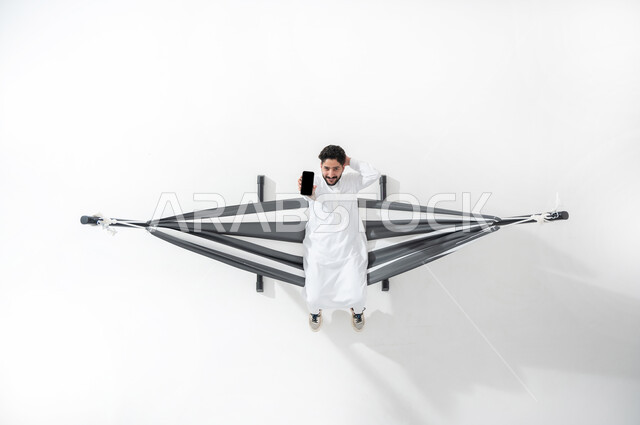 استخدام الأجهزة التقنية الحديثة , بورتريه علوي عامودي لشاب عربي خليجي سعودي  يرتدي الثوب التقليدي يجلس على أرجوحة قماشية معلقة يحمل بيده الهاتف المحمول , النظر إلى الكاميرا بإيماءات السعادة و السرور عرض شاشة سوداء فارغة عبر الجوال , خلفية بيضاء