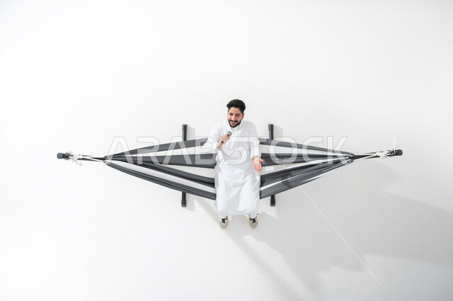أنشطة ترفيهية مسلية، أداء المواهب الصوتية، بورتريه علوي عامودي لرجل عربي خليجي سعودي يرتدي الثوب السعودي التقليدي، يجلس على أرجوحة قماشية، يقوم بالغناء باستخدام المايكروفون، النظر للكاميرا بايماءات الاندماج والتفاعل، تسجيل مقطع صوتي غنائي، خلفية بيضاء
