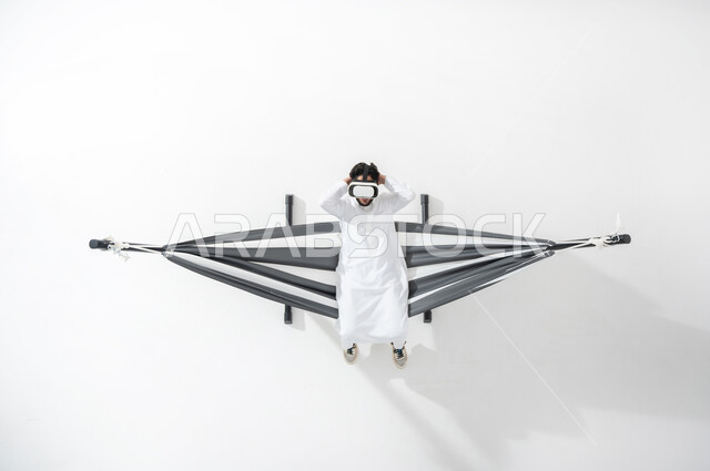 التفاعل ومحاكاة العالم الخيالي،بورتريه علوي عامودي لرجل عربي سعودي خليجي يرتدي الثوب التقليدي يجلس على أرجوحة قماشية معلقة، يستمتع بنظارة الواقع الافتراضي، الجلوس على سرير قماشي ومشاهدة الأفلام عبر نظارات ار ثلاثية الأبعاد، الأجهزة التكنولوجية والتقنيات الحديثة، خلفية بيضاء