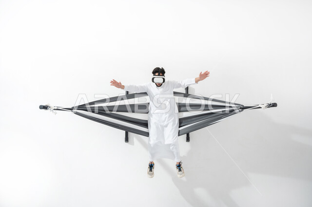 التفاعل ومحاكاة العالم الخيالي،بورتريه علوي عامودي لرجل عربي سعودي خليجي يرتدي الثوب التقليدي يجلس على أرجوحة قماشية معلقة، يستمتع بنظارة الواقع الافتراضي، الجلوس على سرير قماشي ومشاهدة الأفلام عبر نظارات ار ثلاثية الأبعاد، الأجهزة التكنولوجية والتقنيات الحديثة، خلفية بيضاء