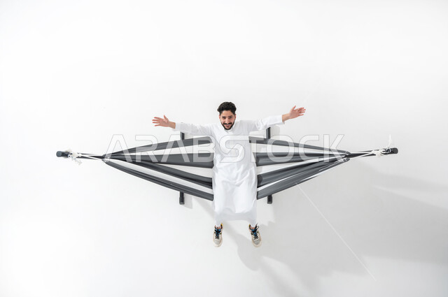 الاستمتاع بوقت الفراغ، بورتريه علوي عامودي لرجل عربي سعودي خليجي يرتدي الثوب التقليدي يجلس على أرجوحة قماشية معلقة، يبسط ذراعيه، الجلوس على سرير قماش، الشعور بالراحة والسكينة، خلفية بيضاء