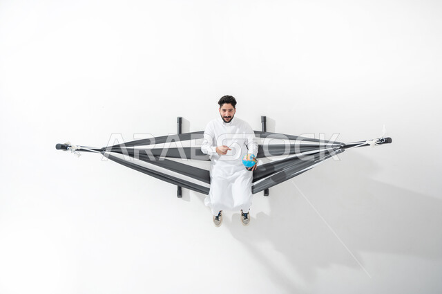 الإشارة بإصبع السبابة إلى مجسم الكرة الأرضية، مفهوم الحفاظ على البيئة في كوكب الارض،التسلية والمرح في أوقات الاستراحة والفراغ، بورتريه علوي عامودي  لشاب عربي سعودي خليجي يرتدي الثوب التقليدي يجلس على أرجوحة قماشية معلقة ينظر إلى الكاميرا ، قضاء الوقت مع الذات، خلفية بيضاء