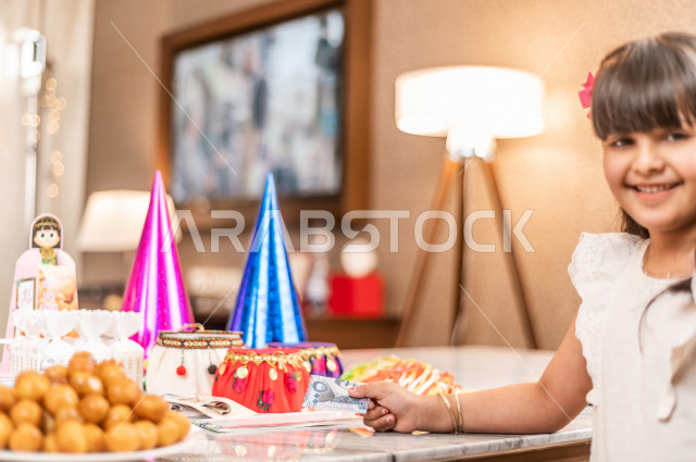 طفلة سعودية خليجية سعيدة ترتدي لباس العيد الانيق ، تشارك الاجواء الاحتفالية الخاصة بعيد الفطر المبارك ، سفرة مليئة بأصناف الحلويات ، الاحتفال بعيد الفطر السعيد ، عيد الفطر المبارك  ،  تقديم العيديات و الحلوى ، القرقيعان