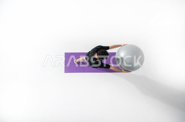 Playing with a balance ball (Pilates), a vertical overhead portrait of a Saudi Arabian Gulf man wearing sports clothing playing with a fitness ball while pushing it with his hands to his left, white background.