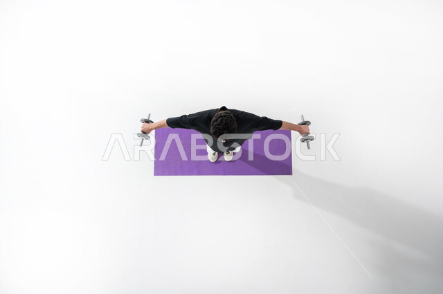 ممارسة التمارين الرياضية من خلال رفع الأثقال الححديدية (الدمبلز)، بورتريه علوي عامودي لرجل عربي خليجي سعودي يرتدي ملابس رياضية ، يقوم ببسط يديه ومحاولة التوازن للمحافظة على اللياقة البدنية