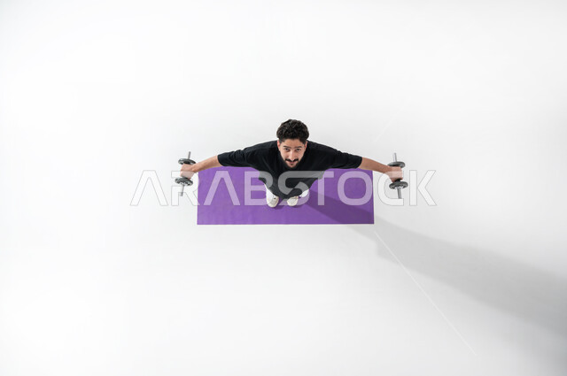 هواية رفع الأثقال الحديدية، بورتريه علوي عامودي لرجل عربي خليجي سعودي يرتدي ملابس رياضية، يقوم بممارسة رفع (الدمبلز)، يبسط يديه  ويرفع رأسه للأعلى، المحافظة على اللياقة البدنية