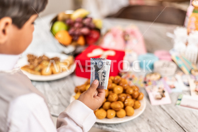 طفل سعودي خليجي سعيد ، يرتدي لباس العيد الانيق ، يأخذ العيدية  ، الاحتفال بعيد الفطر السعيد ، عيد الفطر المبارك  ،  تقديم العيديات و الحلوى ، القرقيعان