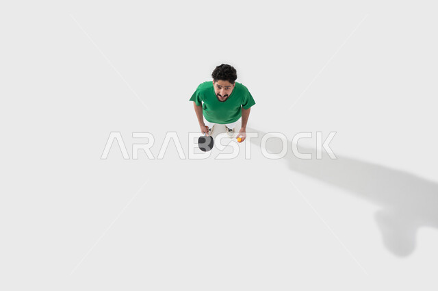 رياضي في الهواء الطلق,  ممارسة رياضات وانشطة بدنية شبابية ممتعة، المحافظة على الرياضة والرشاقة الجسدية، التركيز باللعب واتقانه، بورتريه للاعب عربي خليجي سعودي يرتدي ملابس رياضية يحمل بيده مضرب وكرة تنس الطاولة، خلفية بيضاء