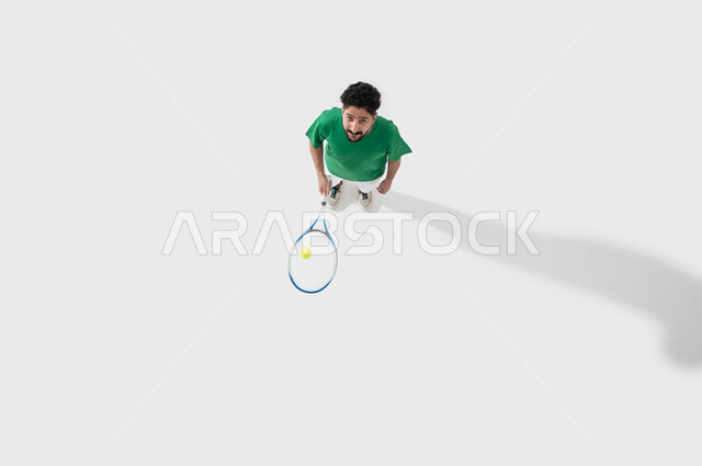 ايماءات التركيز والاندماج في التمرين، المحافظة على الصحة والرشاقة الجسدية، اللعب بسرعة وتركيز، ممارسة رياضات وانشطة بدنية ممتعة، بورتريه لشاب عربي خليجي سعودي يرتدي ملابس رياضية يحمل بيده مضرب تنس، خلفية بيضاء