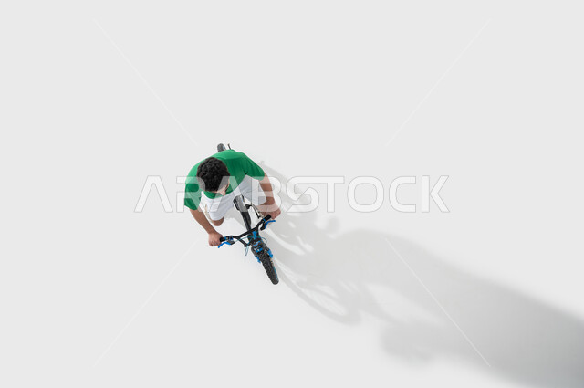 Participating in bicycle racing, practicing the hobby of riding bicycles, physical activity to maintain physical fitness, a vertical overhead portrait of a young Saudi Gulf Arab man riding a bicycle, spending time learning fun and entertaining recreational activities, white background