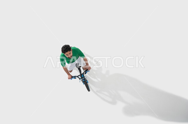 Physical activity to maintain physical fitness, participating in bicycle racing, practicing the hobby of riding bicycles, a vertical overhead portrait of a young Saudi Gulf Arab man riding a bicycle, spending time learning fun and entertaining recreational activities, white background.