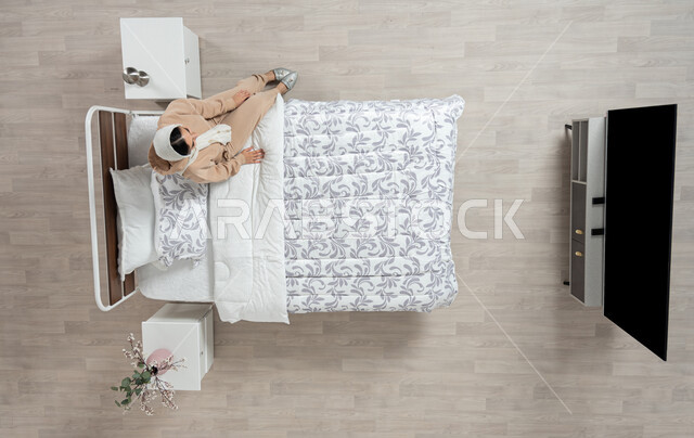 التركيز والتفكير، الشروود والسرحان، بورتريه عامودي علوي لأمرأة عربية خليجية سعودية محجبه تجلس على سريرها بايماءات التمعن في اتخاذ قرارت وحل المشكلات، التفكير بالمستقبل، التعب والارهاق، خلفية بيج