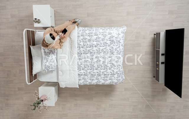 عرض خدمات وتطبيقات الهاتف الجوال، بورتريه عامودي علوي لإمرأة عربية خليجية سعودية محجبه تجلس على سرير مريح تمسك بيدها الهاتف المحمول بشاشة سوداء فارغة، التسويق الإلكتروني عبر الانترنت، التواصل الاجتماعي والدردشه اون لاين، خلفية بيج