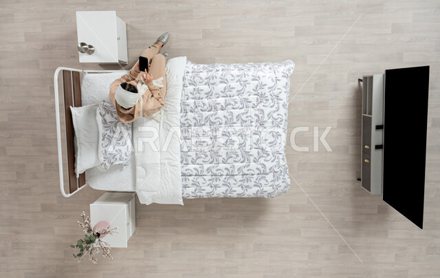 عرض خدمات وتطبيقات الهاتف الجوال، بورتريه عامودي علوي لإمرأة عربية خليجية سعودية محجبه تجلس على سرير مريح تمسك بيدها الهاتف المحمول بشاشة سوداء فارغة، التسويق الإلكتروني عبر الانترنت، التواصل الاجتماعي والدردشه اون لاين، خلفية بيج