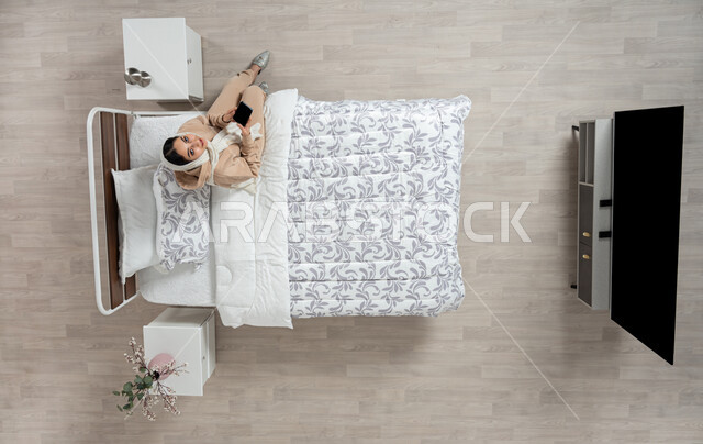 عرض خدمات وتطبيقات الهاتف الجوال، بورتريه عامودي علوي لإمرأة عربية خليجية سعودية محجبه تجلس على سرير مريح تمسك بيدها الهاتف المحمول بشاشة سوداء فارغة، التسويق الإلكتروني عبر الانترنت، التواصل الاجتماعي والدردشه اون لاين، خلفية بيج