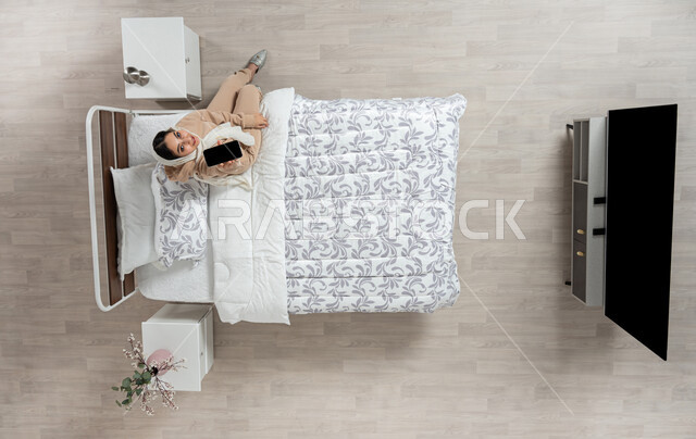 عرض خدمات وتطبيقات الهاتف الجوال، بورتريه عامودي علوي لإمرأة عربية خليجية سعودية محجبه تجلس على سرير مريح تمسك بيدها الهاتف المحمول بشاشة سوداء فارغة، التسويق الإلكتروني عبر الانترنت، التواصل الاجتماعي والدردشه اون لاين، خلفية بيج