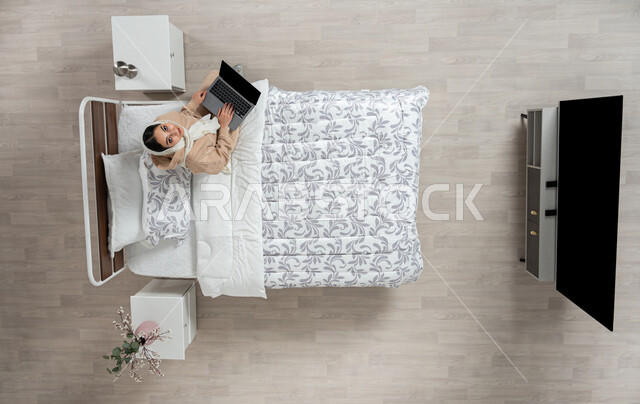 Expressions and gestures of integration and concentration to accomplish tasks and work, using modern and advanced technological devices, an overhead vertical portrait of a smiling Saudi Arabian Gulf woman wearing the hijab sitting on her bed in the bedroom working on her laptop, beige background