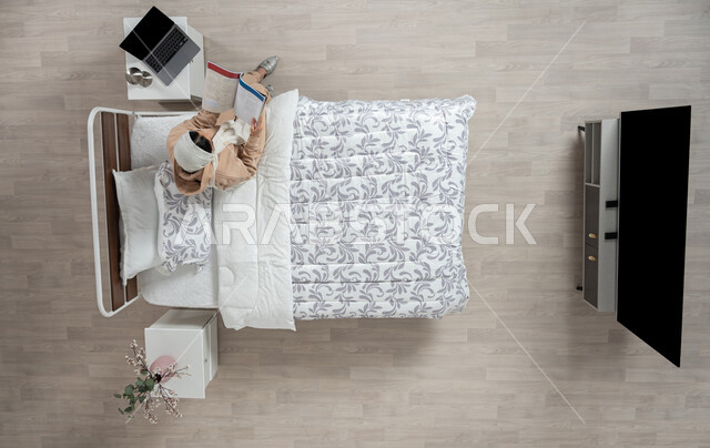 The hobby of reading and exploring, developing and enhancing general information, an overhead vertical portrait of a veiled Saudi Arabian Gulf woman, smiling and wearing casual clothes, sitting on her bed in the bedroom holding a book, spending an enjoyable time reading, interest in science and culture, beige background