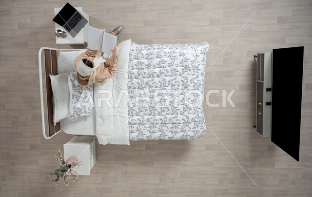 The hobby of reading and exploring, developing and enhancing general information, an overhead vertical portrait of a veiled Saudi Arabian Gulf woman, smiling and wearing casual clothes, sitting on her bed in the bedroom holding a book, spending an enjoyable time reading, interest in science and culture, beige background
