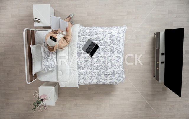 The hobby of reading and exploring, developing and enhancing general information, an overhead vertical portrait of a veiled Saudi Arabian Gulf woman, smiling and wearing casual clothes, sitting on her bed in the bedroom holding a book, spending an enjoyable time reading, interest in science and culture, beige background